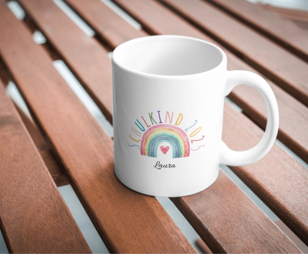 Personalisierte Tasse „Schulkind 2025“ mit Regenbogen und Herz, Name „Laura“, fotografiert auf einem Holztisch.